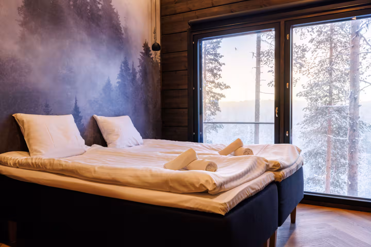 Wide bed beside a tall window looking out over snow-dusted trees at dusk