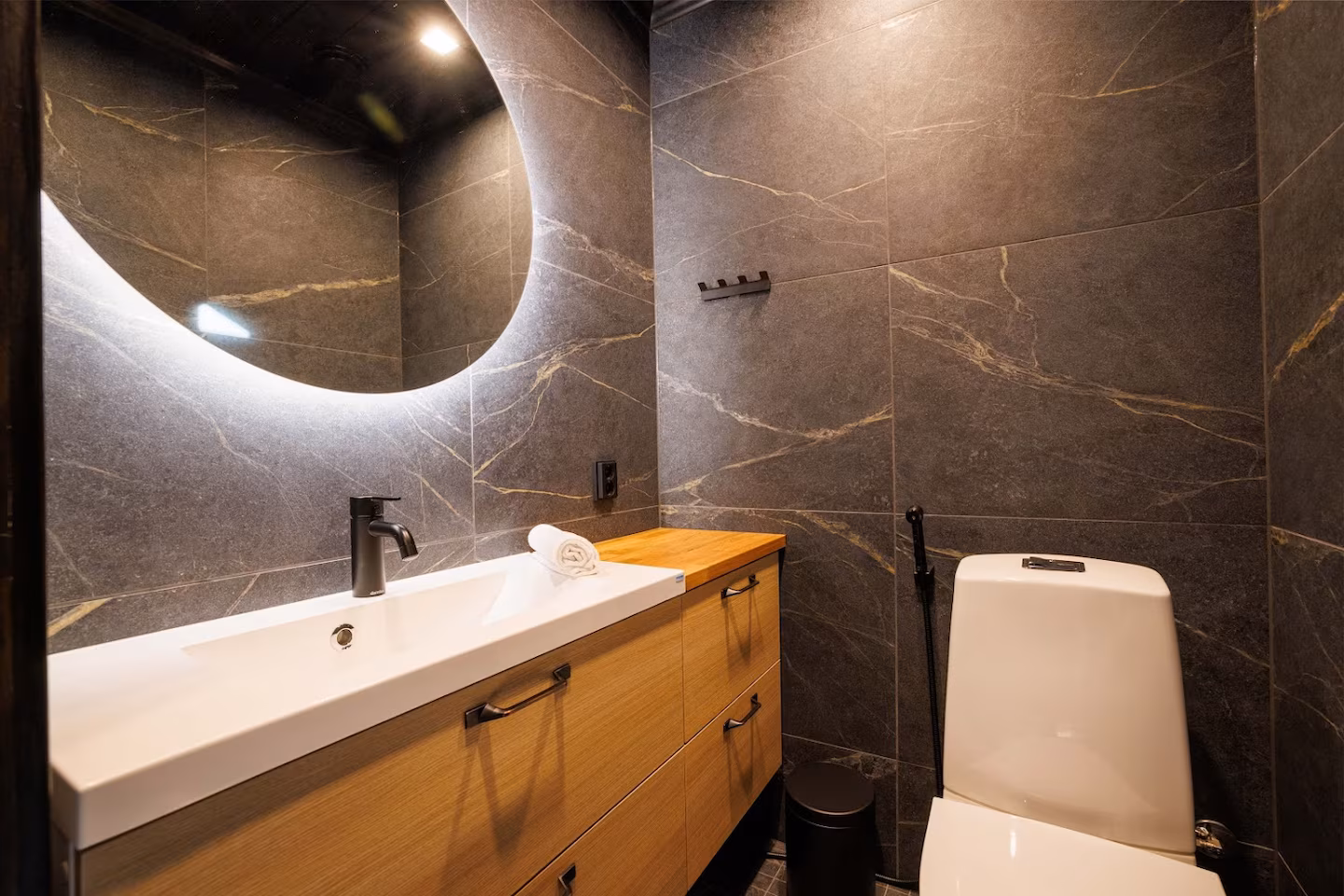 Stone-tiled washroom with a backlit round mirror and oak vanity