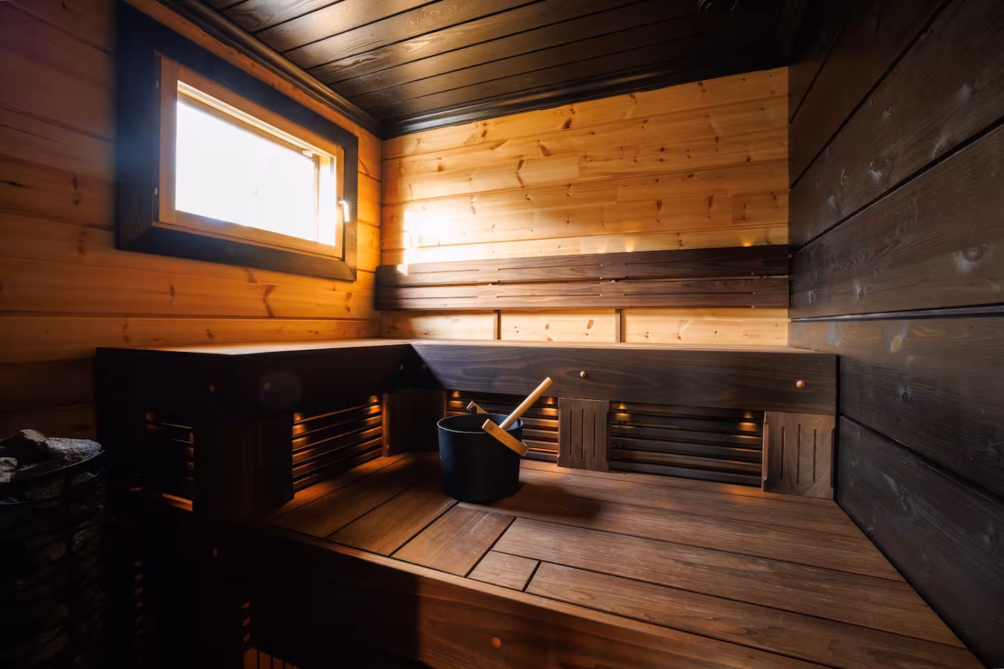 Dark timber sauna with tiered benches, a small window and a black water pail