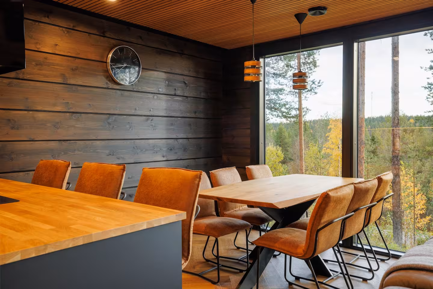 Long dining table with tan chairs beside tall windows onto autumn forest